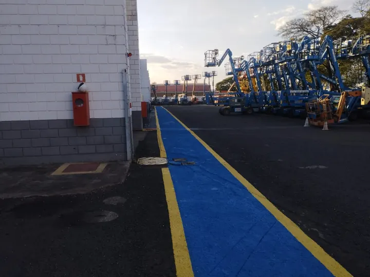 Caminho seguro para pedestres pintado de azul com bordas amarelas em pátio industrial, ao lado de plataformas elevatórias