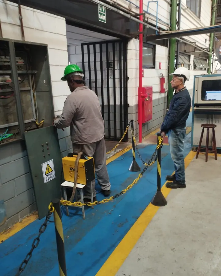Dois técnicos da JW de capacete trabalhando em painel elétrico industrial, com a área isolada por correntes