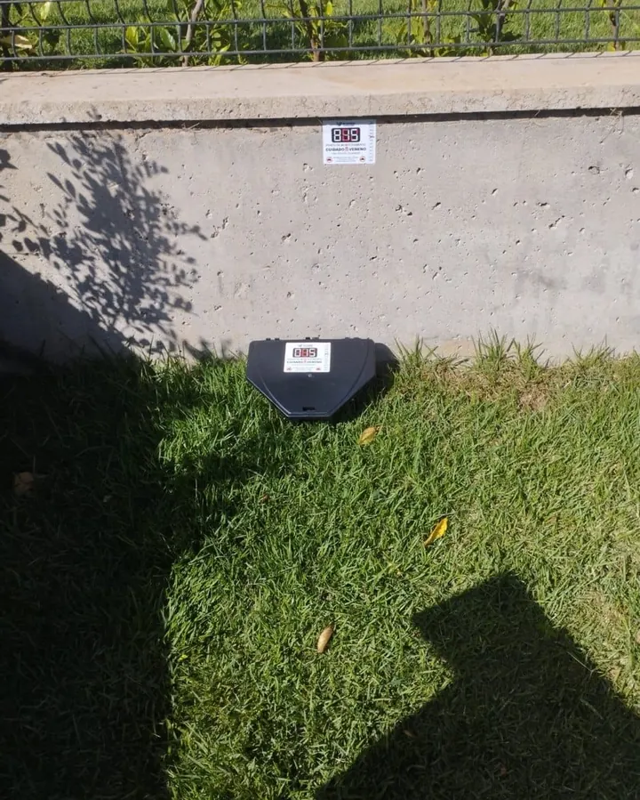Porta-isca para roedores com etiqueta de identificação instalado na base do muro, junto ao gramado